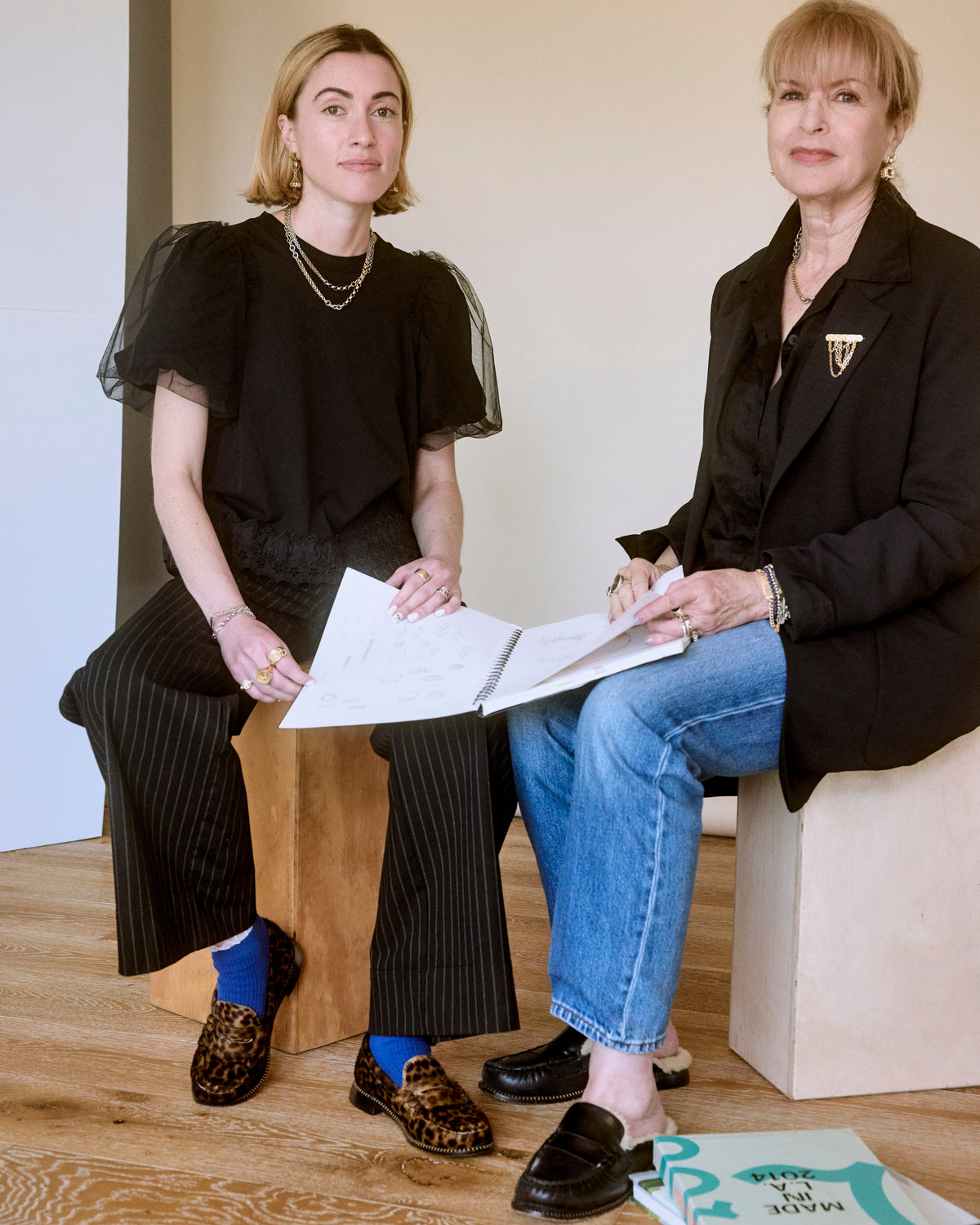 Two women, Jenny and Lily Monbouquette, founders of Monbouquette Jewelry, sitting on stools, one holding a book, in a minimal indoor setting.